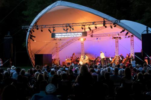 An outdoor concert with a large audience under a canopy, featuring musicians and a screen displaying lyrics.
