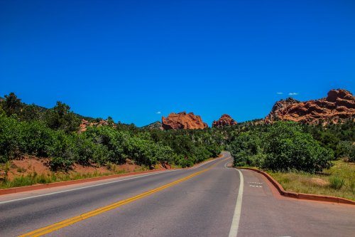 A winding road leads through lush greenery with red rock formations under a clear blue sky.