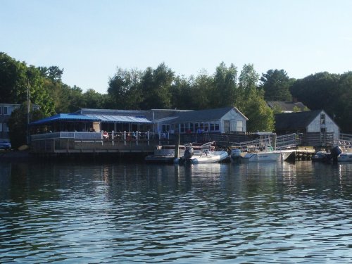 A waterfront restaurant with outdoor seating, surrounded by trees and boats docked in calm water.