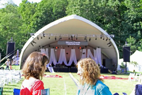 Two women chat in front of a large outdoor stage set up for an event, surrounded by chairs and greenery.