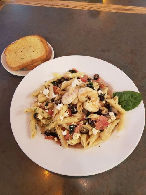 A plate of penne pasta with shrimp, olives, tomatoes, and feta cheese, served with a slice of garlic bread.