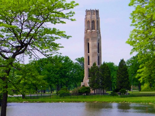 A tall stone tower surrounded by lush green trees and a calm lake under a clear blue sky.