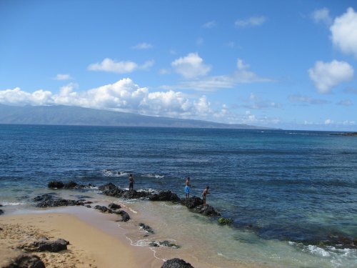 Two people stand on rocks by the shore, with a clear blue sky and mountains in the background.