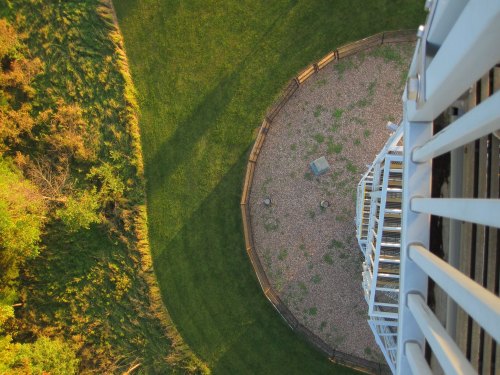 Aerial view of a circular area with grass and gravel, seen from the top of a white tower. Shadows cast on the ground.