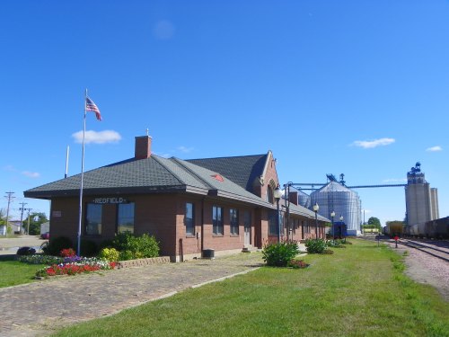 Historic brick train station with American flag, surrounded by flowers and grain silos under a clear blue sky.
