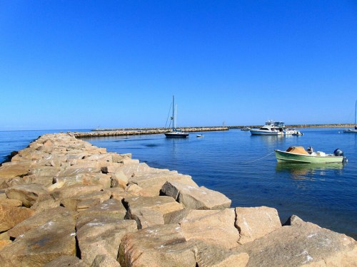 A rocky jetty extends into calm blue waters, with boats moored nearby under a clear blue sky.