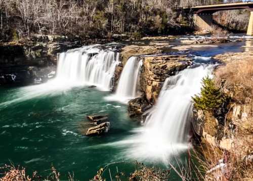 A scenic view of cascading waterfalls with a bridge overhead, surrounded by trees and rocky terrain.