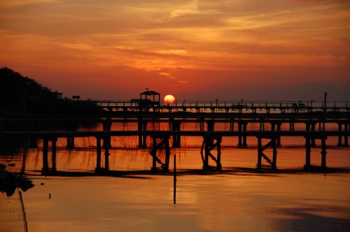 A serene sunset over a calm lake, with silhouettes of a pier and a gazebo against vibrant orange and yellow skies.