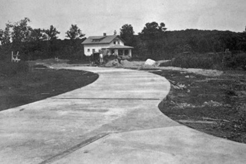 A winding concrete driveway leads to a house surrounded by trees and construction materials.