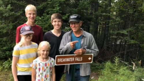A group of five people, including children and an adult, pose by a sign for Breakwater Falls in a forested area.