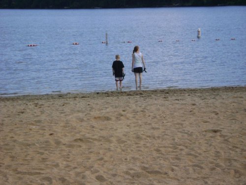 Two children walk hand in hand along the sandy shore of a lake, with calm water and buoys in the background.