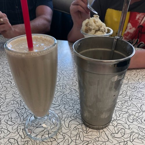 A milkshake in a glass with a straw next to a metal cup and a person holding a bowl of ice cream.