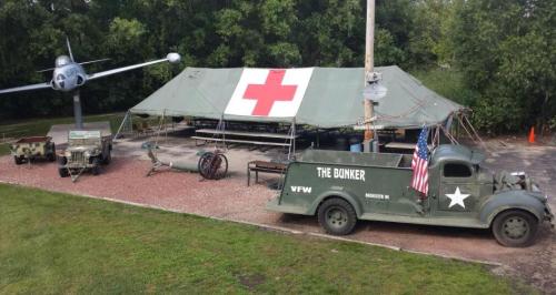 A military-themed display featuring a vintage truck, a medical tent with a red cross, and a plane model nearby.