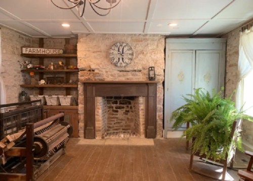 Cozy farmhouse interior featuring a stone fireplace, wooden shelves, and a large clock, with a fern and vintage decor.