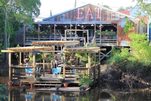 Rustic restaurant with "EAT" sign, surrounded by greenery and water, featuring a wooden deck and outdoor seating.