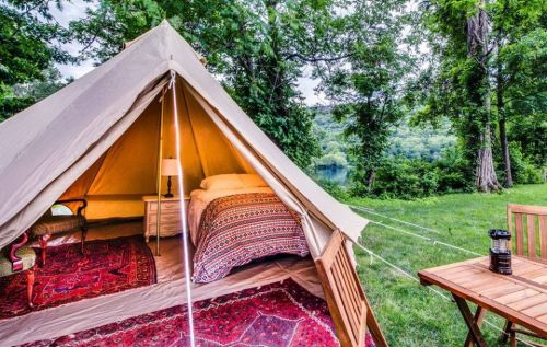 A cozy tent with a bed and rug, set in a scenic outdoor area with trees and a river in the background.