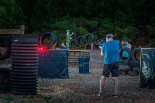 Two people in tactical gear aim laser guns at targets in a wooded outdoor shooting range, surrounded by tires and barriers.