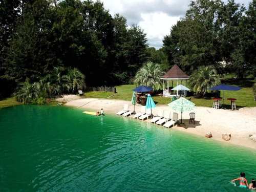 A serene lakeside scene with lounge chairs, umbrellas, and people enjoying the water surrounded by lush greenery.