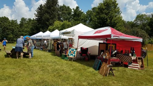 A vibrant outdoor market with tents, vendors, and visitors browsing handmade goods on a sunny day.