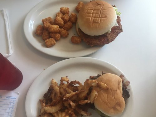 A plate with a crispy chicken sandwich, lettuce, and tater tots, alongside another plate with a burger and onion rings.