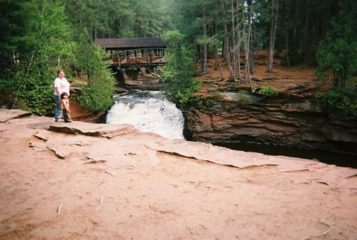 A person stands on a rocky ledge by a waterfall, with a bridge and lush trees in the background.