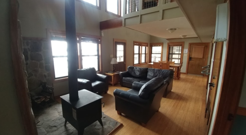 Cozy living room with a stone fireplace, leather sofas, wooden dining table, and large windows letting in natural light.