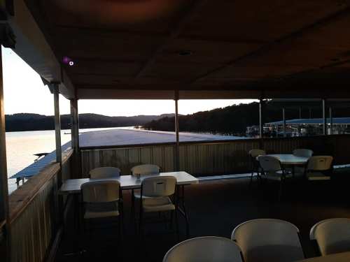 View from a lakeside restaurant at sunset, with tables and chairs overlooking the water and hills in the distance.