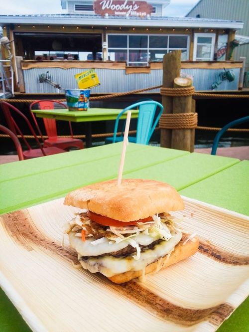 A close-up of a burger on a wooden plate, with a colorful outdoor dining area and a food truck in the background.