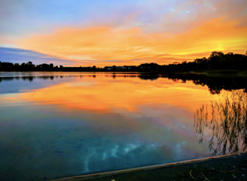 A serene lake at sunset, reflecting vibrant orange and blue hues in the water, surrounded by trees and a peaceful sky.