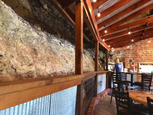 Interior of a rustic restaurant featuring exposed rock walls, wooden beams, and a bar area with a person preparing drinks.
