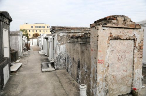 A narrow path between weathered tombs in a cemetery, with a building visible in the background under a cloudy sky.