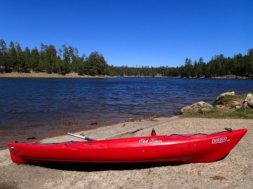 A red kayak rests on a sandy shore by a calm lake, surrounded by green trees and a clear blue sky.