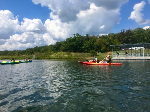 Two people kayaking on a calm lake with green trees and fluffy clouds in the background.