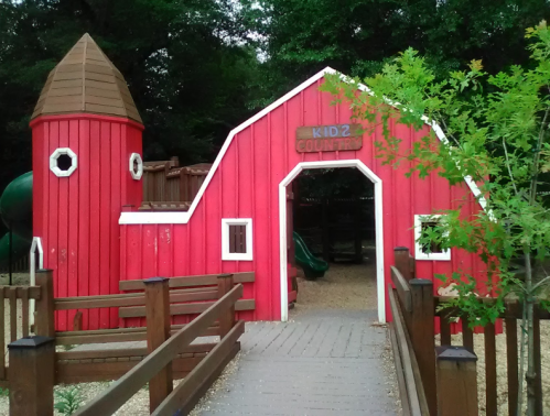 A red play barn with a tower, featuring a sign that reads "KIDZ COUNTRY," surrounded by wooden fencing and greenery.