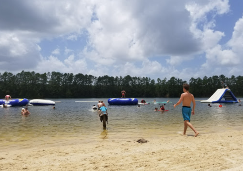 A sandy beach with people swimming and playing on inflatable structures in a lake under a partly cloudy sky.