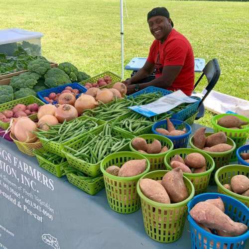 A smiling vendor sits behind a table filled with fresh vegetables and baskets of sweet potatoes at a farmers market.