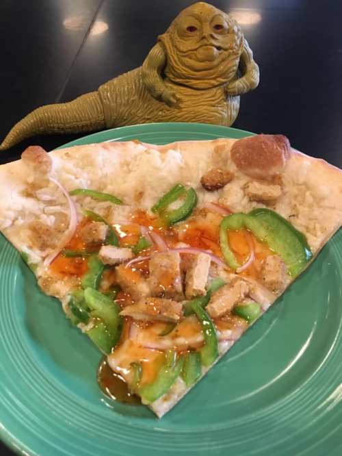 A slice of pizza topped with green peppers, chicken, and sauce, next to a Jabba the Hutt figurine.
