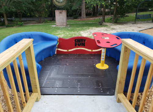 A colorful playground seating area with blue curved benches and a red table, surrounded by trees and a sandy area.