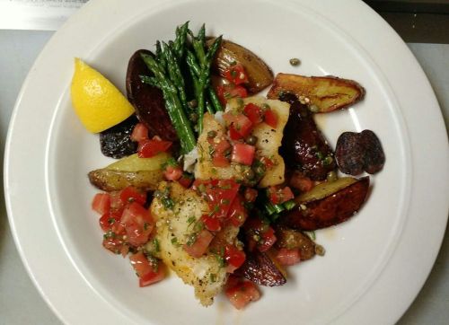 A plate of grilled fish topped with diced tomatoes, served with roasted potatoes, asparagus, and a lemon wedge.