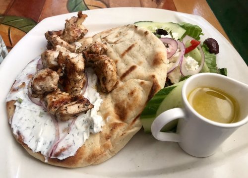A plate featuring grilled chicken on flatbread with yogurt sauce, a side salad, and a small cup of olive oil.