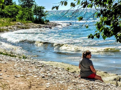 A person sits on a sandy shore, overlooking gentle waves and a sparkling lake under a clear blue sky.