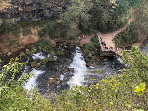 Aerial view of a river with rapids, surrounded by greenery and a wooden viewing platform with people nearby.