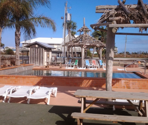 A sunny pool area with lounge chairs, a thatched gazebo, and palm trees in the background.