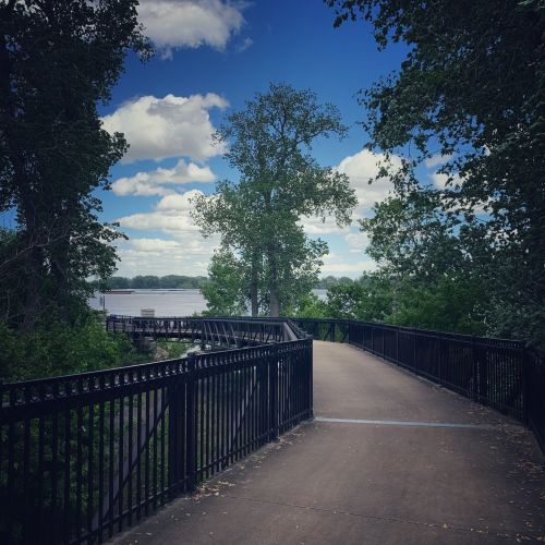 A scenic pathway lined with trees, leading towards a river under a partly cloudy sky.
