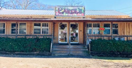 A wooden building with a sign reading "Carol's" and an "Open" neon sign, surrounded by greenery.