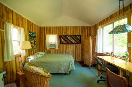 Cozy wooden interior of a room featuring a bed, desk, chair, and large windows with natural light.