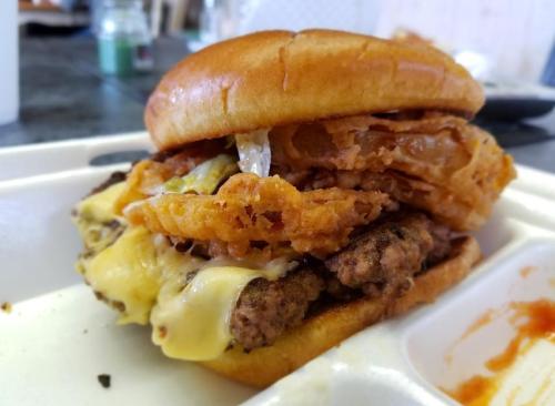 A close-up of a burger with two beef patties, melted cheese, crispy onion rings, and a soft bun on a takeout container.