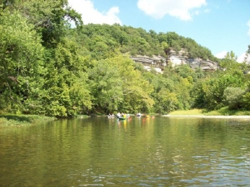 A serene river scene with kayakers paddling among lush green trees and rocky cliffs under a blue sky.