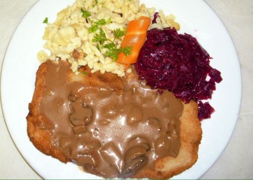 A plate featuring breaded meat with mushroom gravy, spätzle, red cabbage, and a carrot garnish.