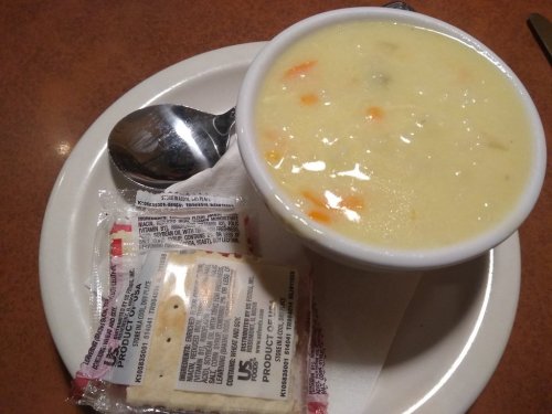 A bowl of creamy soup with vegetables, accompanied by a packet of crackers and a spoon on a plate.